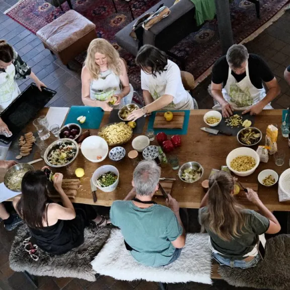 Gruppe von acht Menschen kocht gemeinsam an einem großen Holztisch in einer Küche.