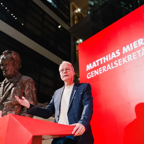Mann spricht an Rednerpult neben einer Bronzestatue, roter Hintergrund mit Schriftzug.