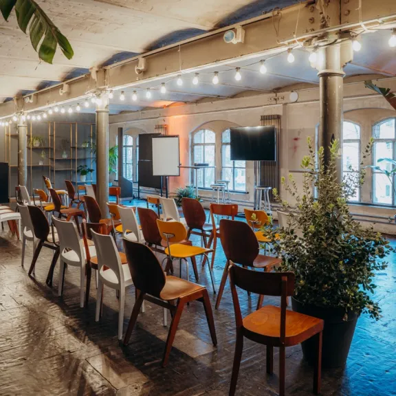 Stuhlreihen in einem hellen, gemütlichen Veranstaltungsraum mit Pflanzen und Lichterketten.