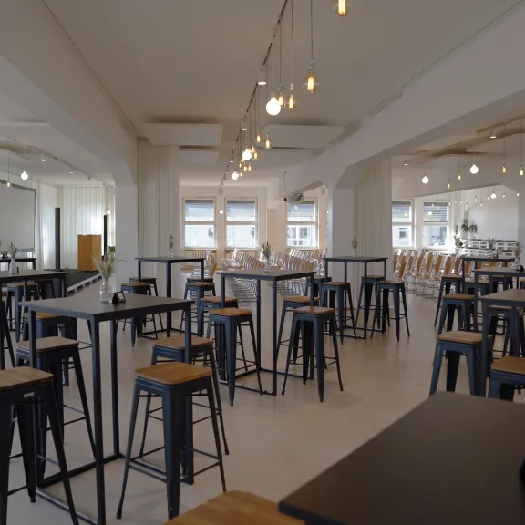 Heller Raum mit vielen hohen Tischen und Hockern, moderne Einrichtung, warme Beleuchtung.