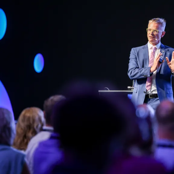 Mann im Anzug spricht vor Publikum auf einer Bühne mit blauem Licht.