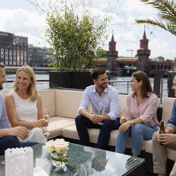 Fünf Menschen sitzen lachend auf einer Terrasse mit Blick auf die Oberbaumbrücke.