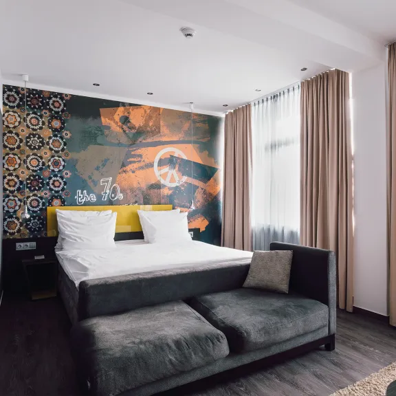 Modernes Hotelzimmer mit Doppelbett, Sofa und bunter Wandkunst, große Fenster mit Vorhängen.