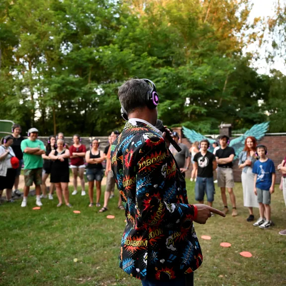 Moderator mit Mikrofon und Kopfhörern leitet ein interaktives Quiz im Freien; die Teilnehmenden stehen im Kreis und tragen kabellose Kopfhörer.