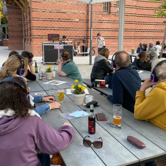 Teilnehmende mit Kopfhörern sitzen an Tischen im Außenbereich und nehmen an einem moderierten Musik-Quiz mit Host teil.