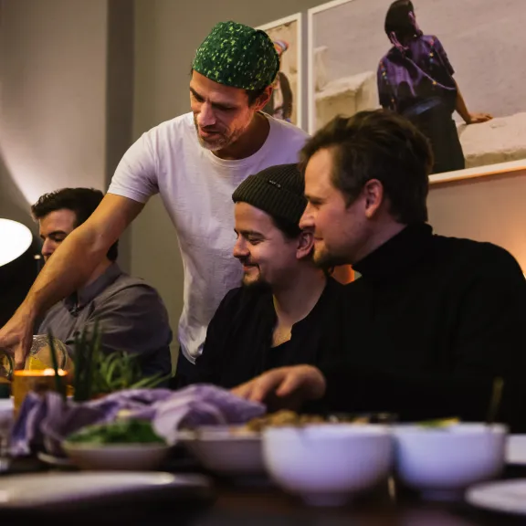 Mehrere Menschen sitzen lachend am gedeckten Tisch, ein Koch bedient sie.