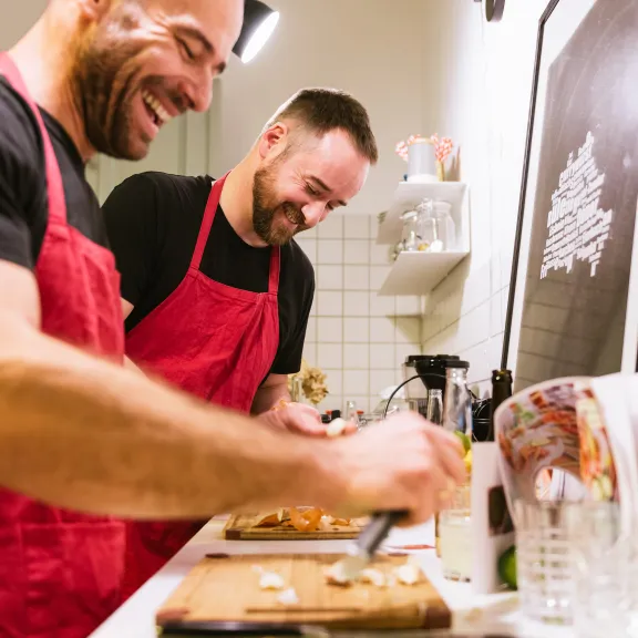 Zwei lachende Männer in roten Schürzen schneiden Gemüse in einer hellen Küche.