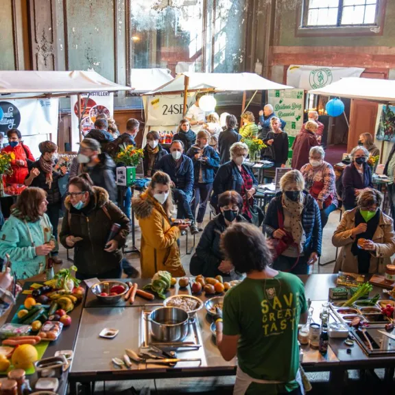 Menschenmenge auf einem Markt mit Ständen, viele tragen Masken und halten Blumen.