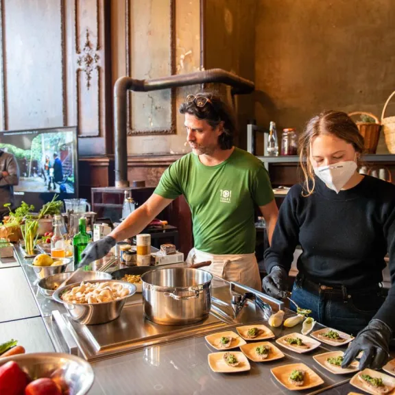Zwei Menschen bereiten Speisen in einer Küche zu, mit frischen Zutaten auf dem Tisch.