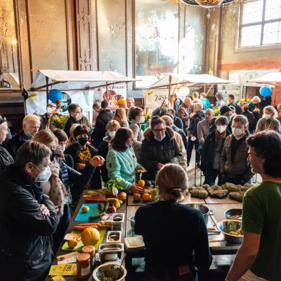 Viele Menschen auf einem gut besuchten Markt in einer Halle mit Ständen und Lebensmitteln.