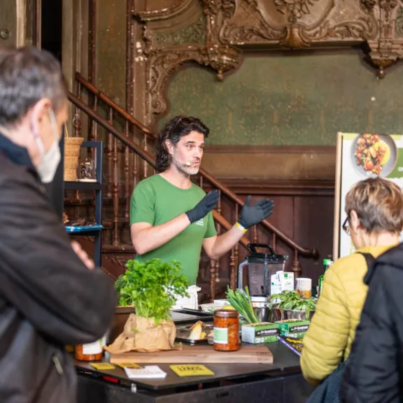 Mann erklärt einer kleinen Gruppe Pflanzen auf einem Marktstand in einem alten Gebäude.