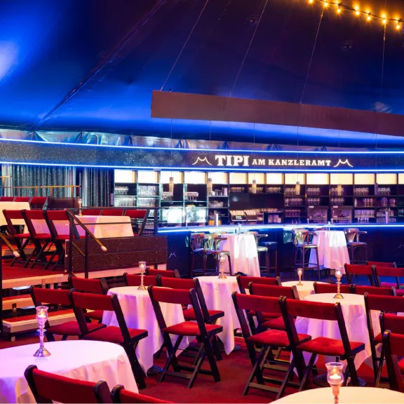 Festlich beleuchteter Veranstaltungsraum mit Tischen, roten Stühlen und einer Bar im Hintergrund.