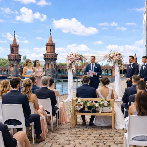 Hochzeitszeremonie im Freien mit Gästen, Blumen und Tafel vor einer Brücke.