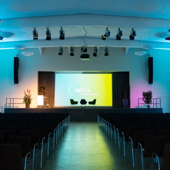 Leerer Konferenzsaal mit beleuchteter Bühne und zwei Sesseln vor einer großen Leinwand