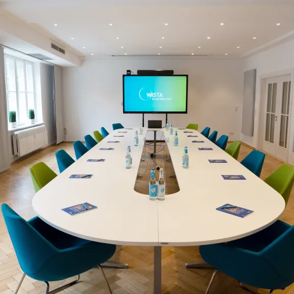 Moderner Konferenzraum mit ovalem Tisch, bunten Stühlen und Bildschirm.