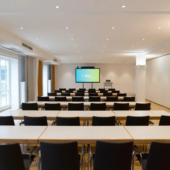 Heller Konferenzraum mit Tischen, Stühlen und einem Bildschirm vorne.