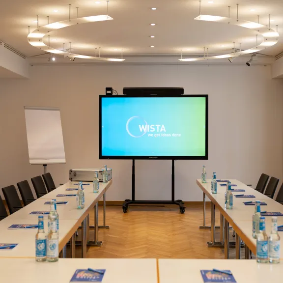 Konferenzraum mit U-förmigem Tisch, Bildschirm, Stühlen und Wasserflaschen.