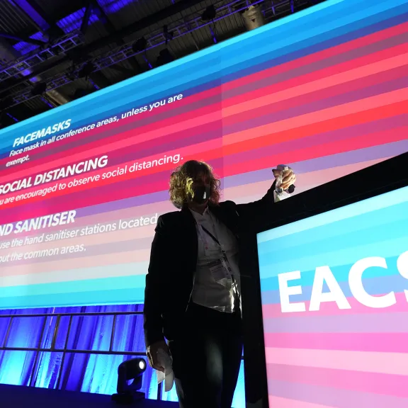 Person mit Mikrofon vor großer, bunter Präsentationswand mit Schriftzug „EACS“.