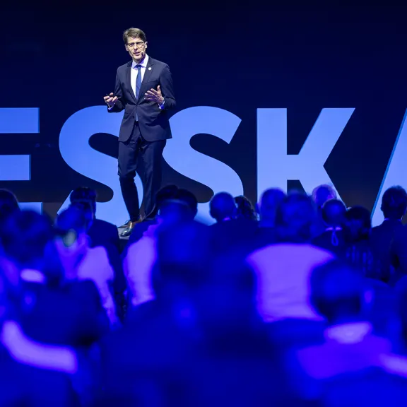Mann im Anzug spricht auf einer Bühne vor großem Publikum, Hintergrund mit ESSKA-Schriftzug.