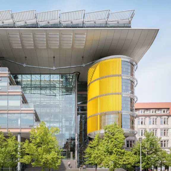 Modernes Gebäude mit Glasfront und gelbem Zylinder, davor grüne Bäume und Gehweg.