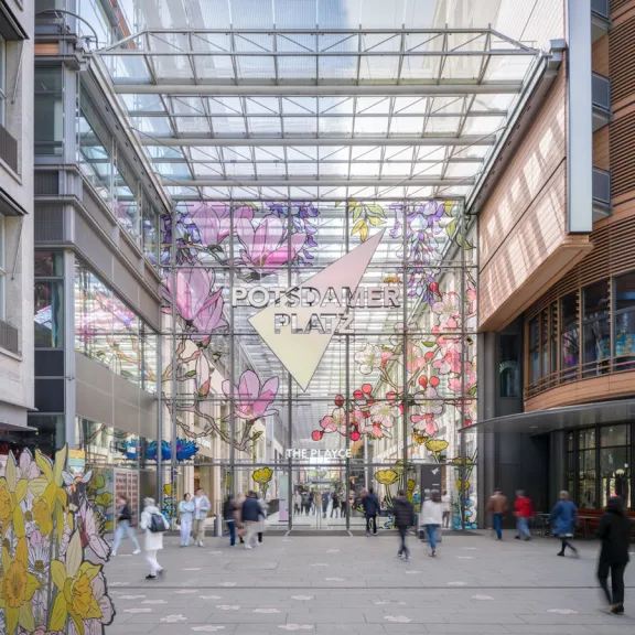 Überdachter Durchgang mit bunten Blumeninstallationen und vorbeigehenden Menschen.
