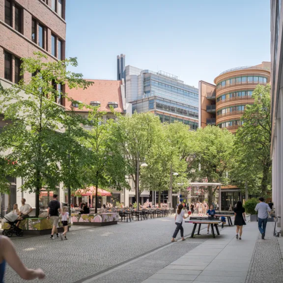 Belebte Fußgängerzone mit Bäumen, modernen Gebäuden und Menschen bei sonnigem Wetter.