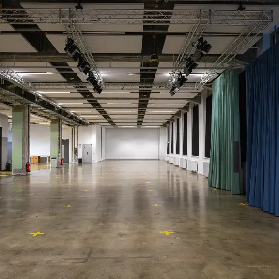Leere Veranstaltungshalle mit Betonboden, Vorhängen und Deckenbeleuchtung.