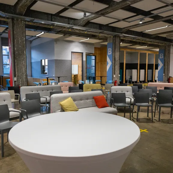 Moderner Veranstaltungsraum mit Stühlen, Sofas und runden Tischen, bunte Kissen.