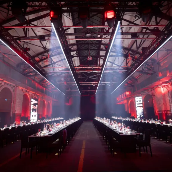 Langer Festsaal mit zwei gedeckten Tafeln, rote Beleuchtung, industrielle Atmosphäre.