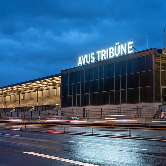 Stadiontribüne bei Dämmerung, beleuchtet, nasse Straße und vorbeifahrende Autos.