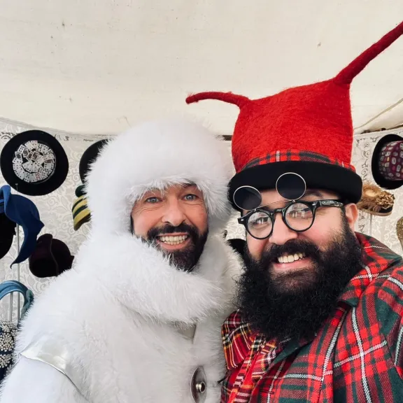 Die beiden HatArts-Inhaber stehen vor einer Wand mit ausgefallenen Hüten; einer trägt ein flauschiges weißes Kostüm, der andere einen hohen roten Hut und eine karierte Jacke.