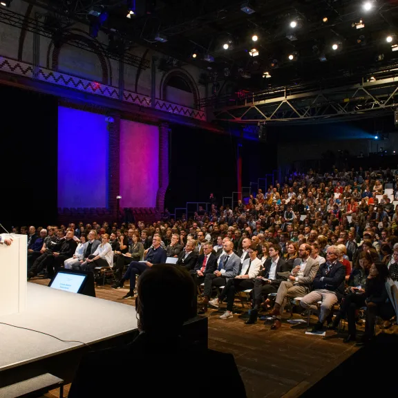 Redner spricht auf großer Bühne vor vollbesetztem Publikum in einem Saal