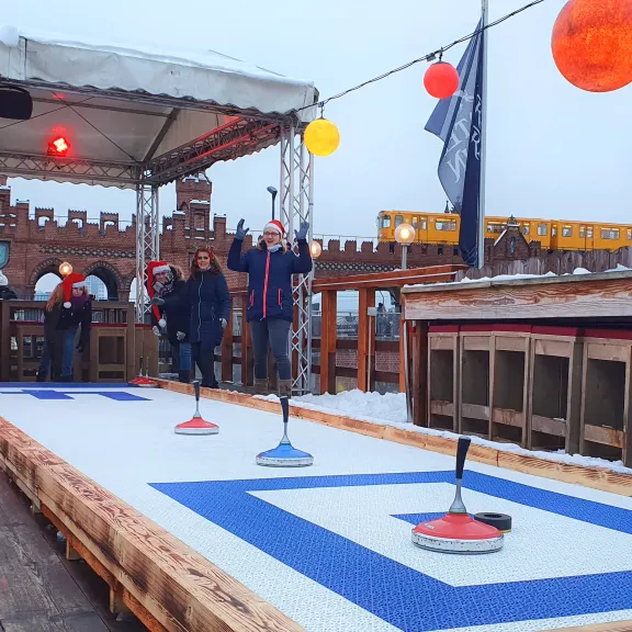 Menschen spielen Eisstockschießen auf einer Bahn im Freien, bunte Lampions hängen darüber.