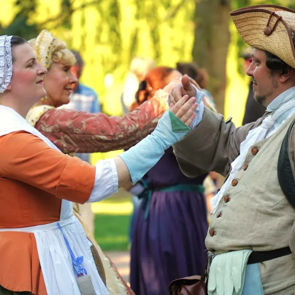 Tanz in historischen Kostümen: Eine Tänzerin und ein Tänzer stehen sich gegenüber und berühren sich mit erhobenen Händen, während weitere Teilnehmende im Hintergrund zuschauen oder mittanzen.