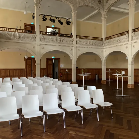 Leerer Saal mit weißen Stühlen, Parkettboden und dekorativen Bögen.