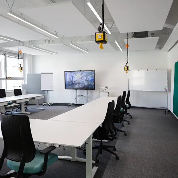 Heller Konferenzraum mit Tischen, Stühlen, Whiteboard und großem Bildschirm.