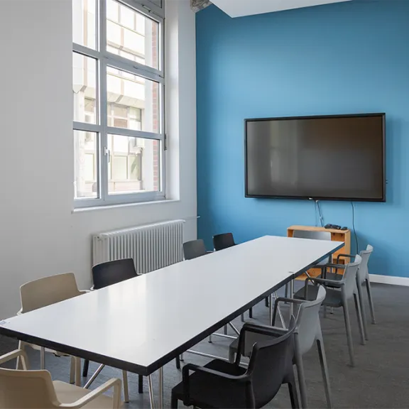 Konferenzraum mit langem Tisch, Stühlen und großem Bildschirm an blauer Wand.