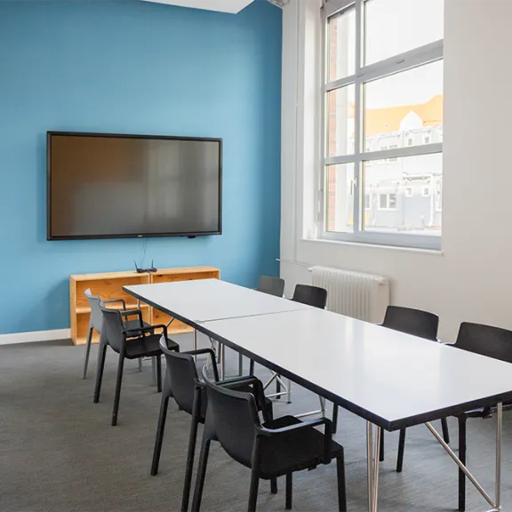 Heller Konferenzraum mit großem Tisch, schwarzen Stühlen und Flachbildfernseher an blauer Wand.