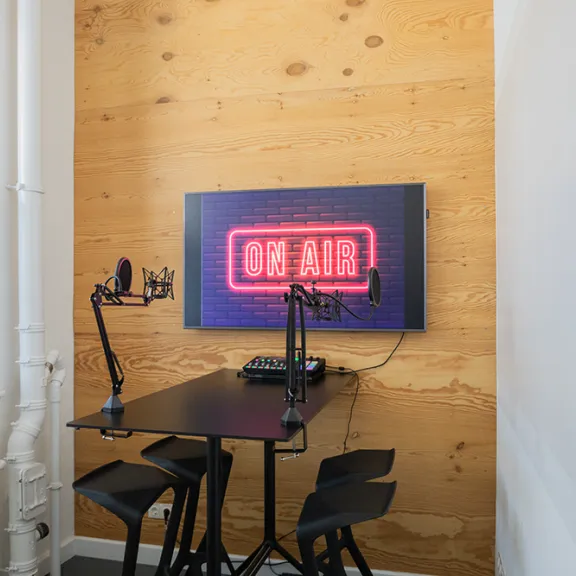 Kleines Podcast-Studio mit Tisch, Mikrofonen und leuchtendem "On Air"-Schild an der Wand.