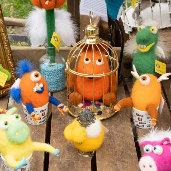 Bunte, handgefertigte Filzfiguren von Königs Filzerei stehen auf einem Tisch; einige sitzen in Tassen oder Glasgefäßen, andere tragen kleine Accessoires.