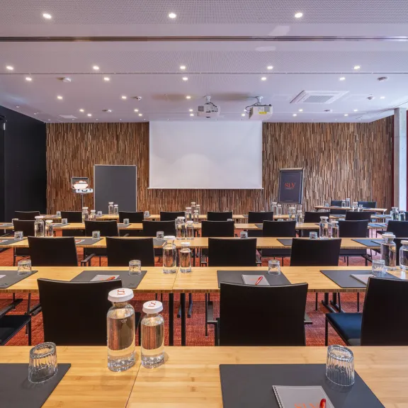 Heller Konferenzraum mit Tischen, Stühlen, Wasserflaschen und Leinwand.