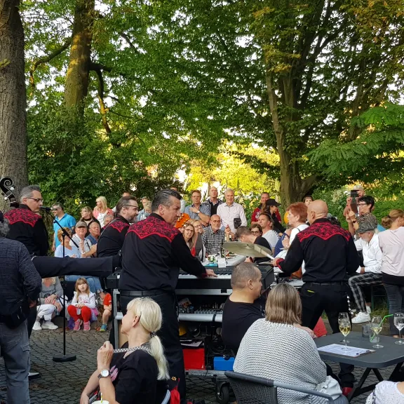 Die Band Krüger Rockt! spielt mobil auf einem Festgelände; Musiker in schwarzen Outfits mit roten Stickereien treten zwischen den Besucherinnen und Besuchern auf, die an Tischen sitzen oder stehen.