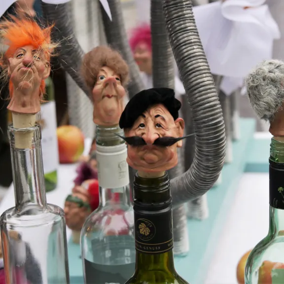 Weinflaschen mit kunstvollen, humorvollen Flaschenverschlüssen in Form von charaktervollen Köpfen stehen auf einem Tisch, umgeben von weiteren Deko‑Objekten