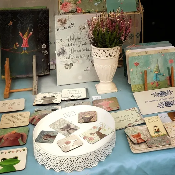 Illustrierte Karten und kleine Deko‑Boards mit Sprüchen und Naturmotiven stehen auf einem Tisch; daneben ein weißer Krug mit Heideblüten