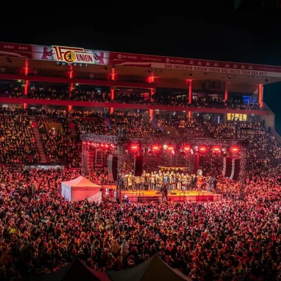 Große Menschenmenge bei einem Konzert im Stadion, Bühne leuchtet rot.