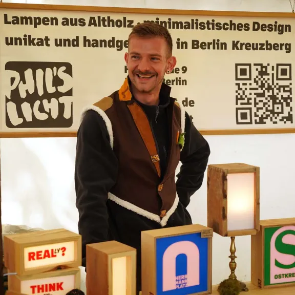 Phil Lehmann steht an seinem Marktstand mit handgefertigten Lampen aus nachhaltigem Altholz; mehrere minimalistische Leuchten mit warmweißem Licht sind ausgestellt.