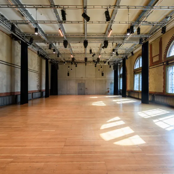 Großer, leerer Saal mit Holzboden und großen Fenstern, Sonnenlicht fällt herein.