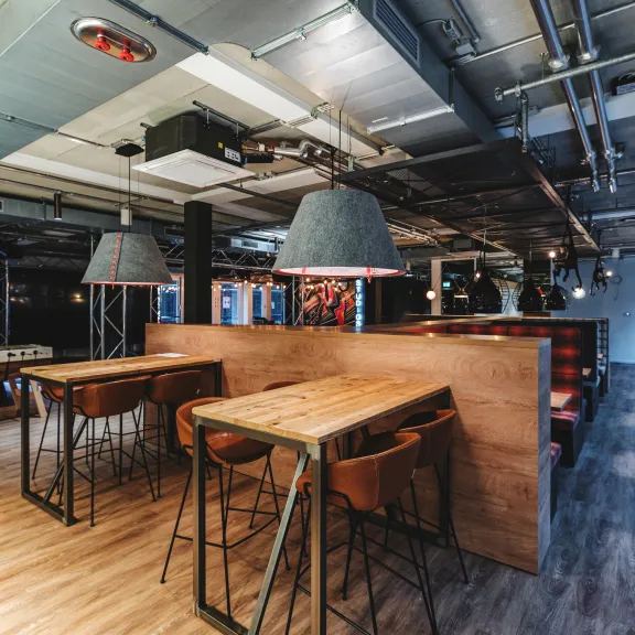 Moderne Veranstaltungsfläche mit Holztischen, Hockern und industriellen Deckenleuchten.