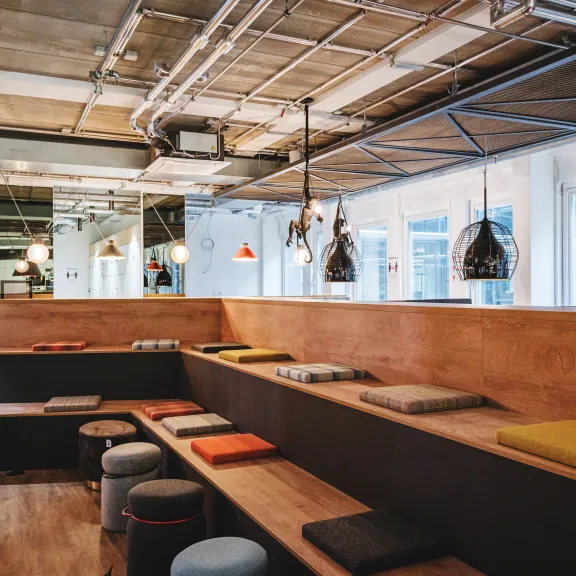 Moderne Veranstaltungsfläche mit Holztribüne, farbigen Sitzkissen und industriellen Lampen.