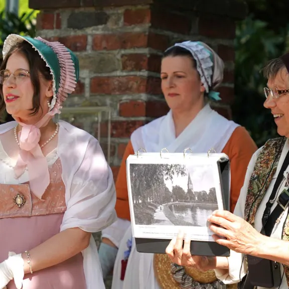 Drei Personen in historischer Kleidung führen eine Stadtteilgruppe; eine von ihnen zeigt ein altes Foto, das die Geschichte von Lichtenrade veranschaulicht.